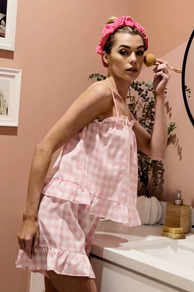 A side view of a woman in a pink gingham lounge set standing at a bathroom counter. She looks into the mirror while adjusting her makeup, with soft pink decor in the background complementing her outfit.