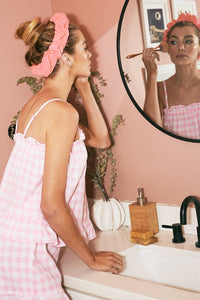 A woman wearing a pink gingham frill lounge set applies makeup with a brush in front of a bathroom mirror. She has a pink scrunchie headband, and the soft lighting enhances the cozy aesthetic.