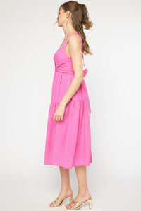 Side view of a model wearing a pink midi dress with a twist front detail and bow tie back, fully lined, made of 100% polyester, against a white background.