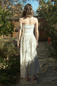 Woman wearing an elegant strapless foldover midi dress in a neutral tone, standing against a rustic wooden backdrop. The dress features a ribbed bodice and flowy skirt, perfect for effortless summer style.







