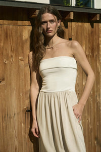 Woman wearing an elegant strapless foldover midi dress in a neutral tone, standing against a rustic wooden backdrop. The dress features a ribbed bodice and flowy skirt, perfect for effortless summer style.







