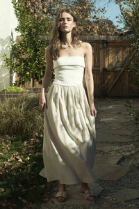 Woman wearing an elegant strapless foldover midi dress in a neutral tone, standing against a rustic wooden backdrop. The dress features a ribbed bodice and flowy skirt, perfect for effortless summer style.







