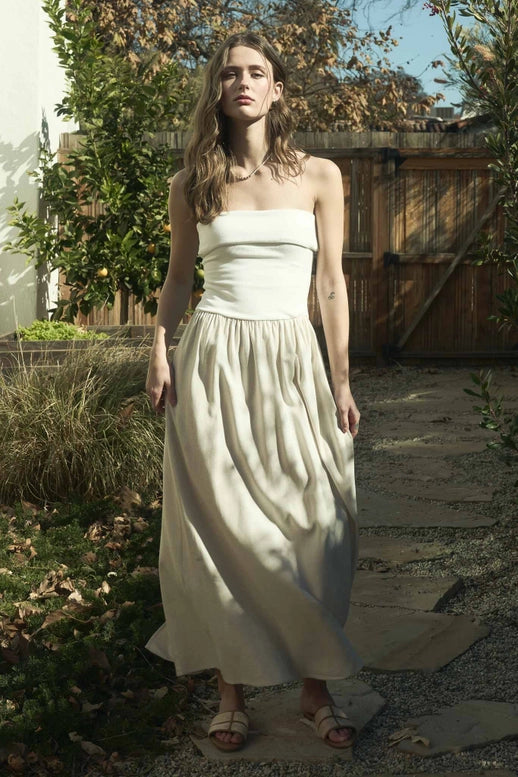 Woman wearing an elegant strapless foldover midi dress in a neutral tone, standing against a rustic wooden backdrop. The dress features a ribbed bodice and flowy skirt, perfect for effortless summer style.







